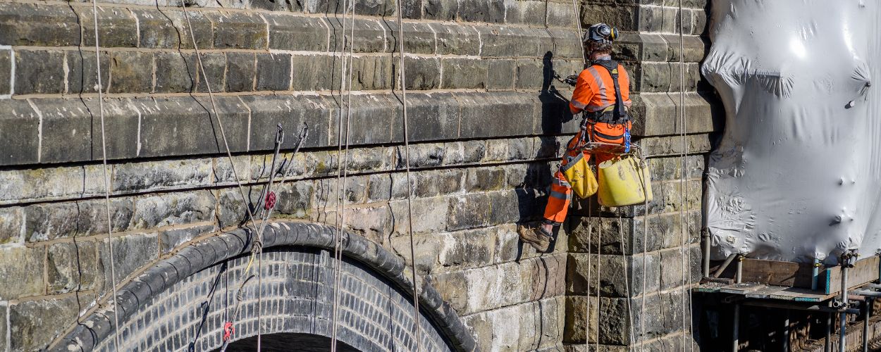 A man wearing a protective suit works on a viaduct stone wall.