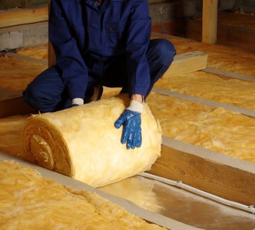 A person in a dark blue boiler suit with blue gloves rolls out a bail of fibreglass insulation in a room.